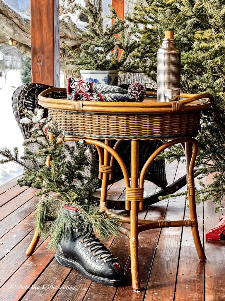 Vintage wicker table on outdoor porch with vintage ski booth, evergreens and old thermos and ski hat.