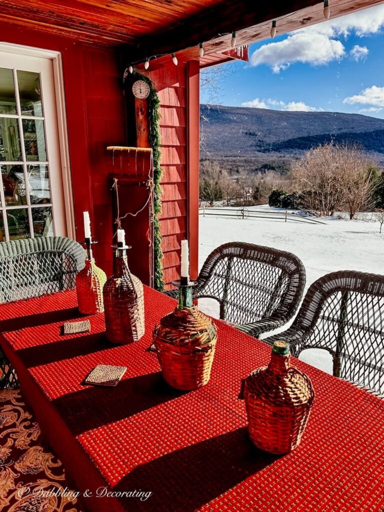 Vintage demijohn bottles lined up on outdoor winter porch table with red tablecloth with vintage wooden sled leaning up against house in the mountains.