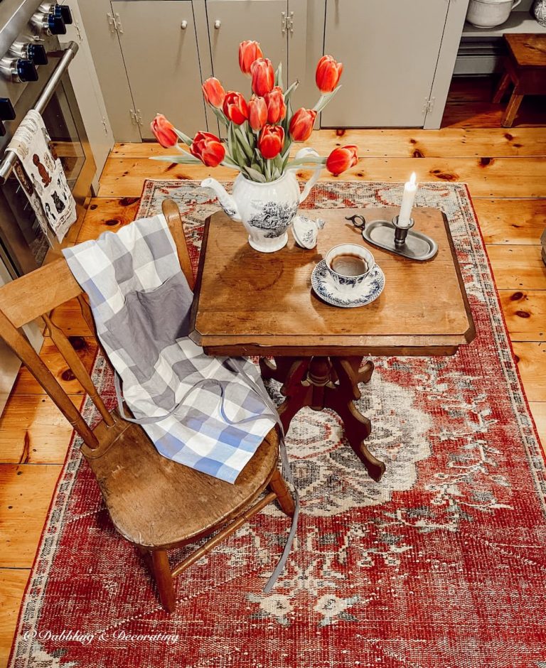 A country primitive style kitchen with wood floors, vintage rug and wooden table and chair with red tulips and pewter candlestick in vintage kitchen decor ideas.