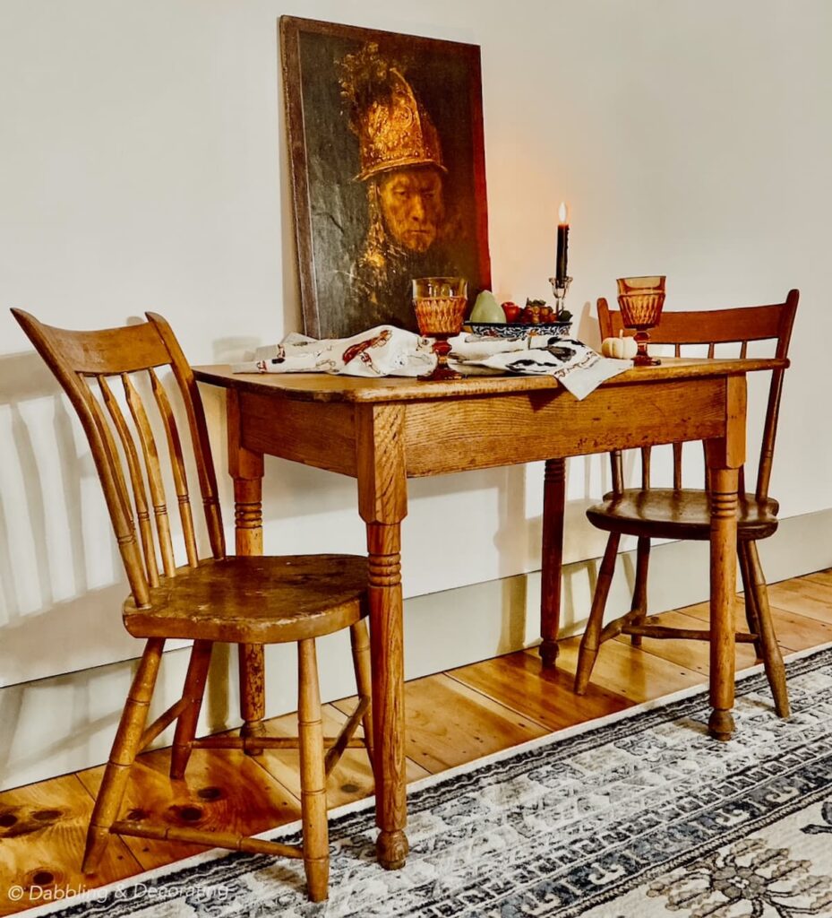 An antique wooden table set for two with two matching antique chairs and a picture of the Man with the Golden Helmet.