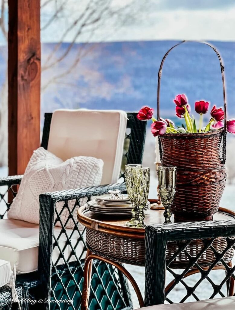 Covered porch furniture ideas with a wicker chair with white cushions and a vintage wicker side table with flowers with mountain views.