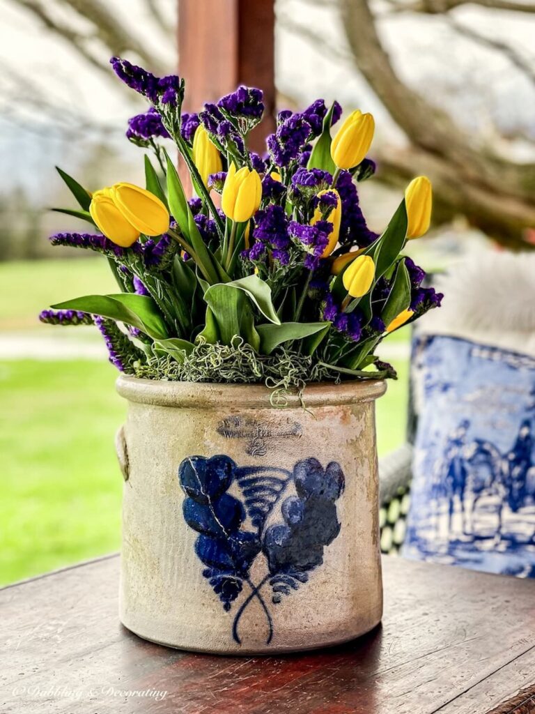 Vintage porch crock filled with yellow and purple flowers on a porch coffee table.