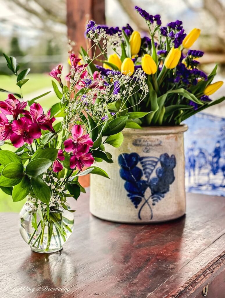 Vintage crock filled with yellow tulips and purple flowers along with a smaller bouquet of pink flowers in a mason jar on covered porch coffee table.