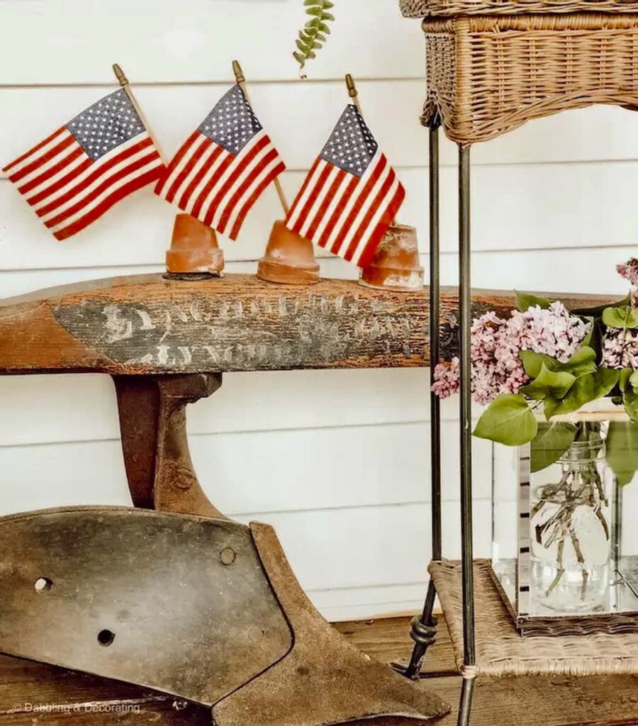 Vintage Americana decor ideas with an old garden tool, America flags in upside down terracotta pots with a bouquet of lilacs on wicker porch table.