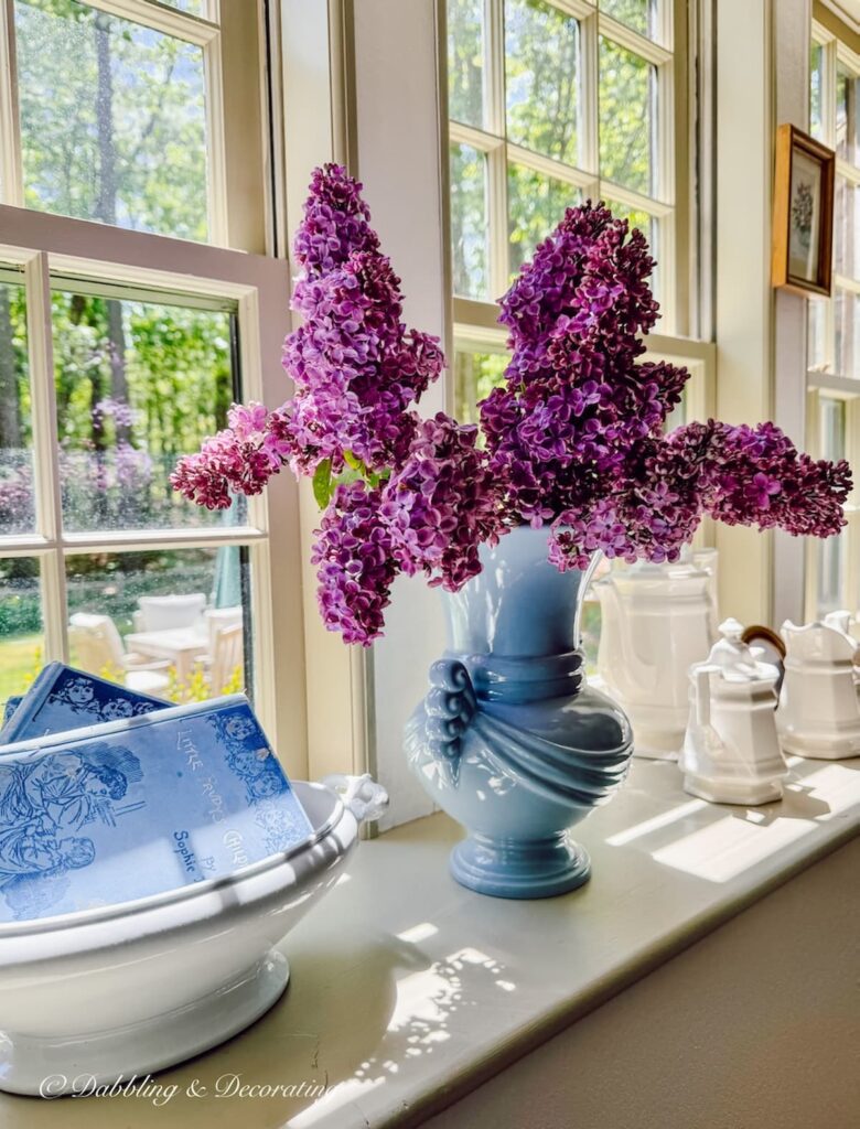 Vintage blue vase filled with lilacs on a large dining room windowsill next to a white ironstone tureen filled with vintage blue books.