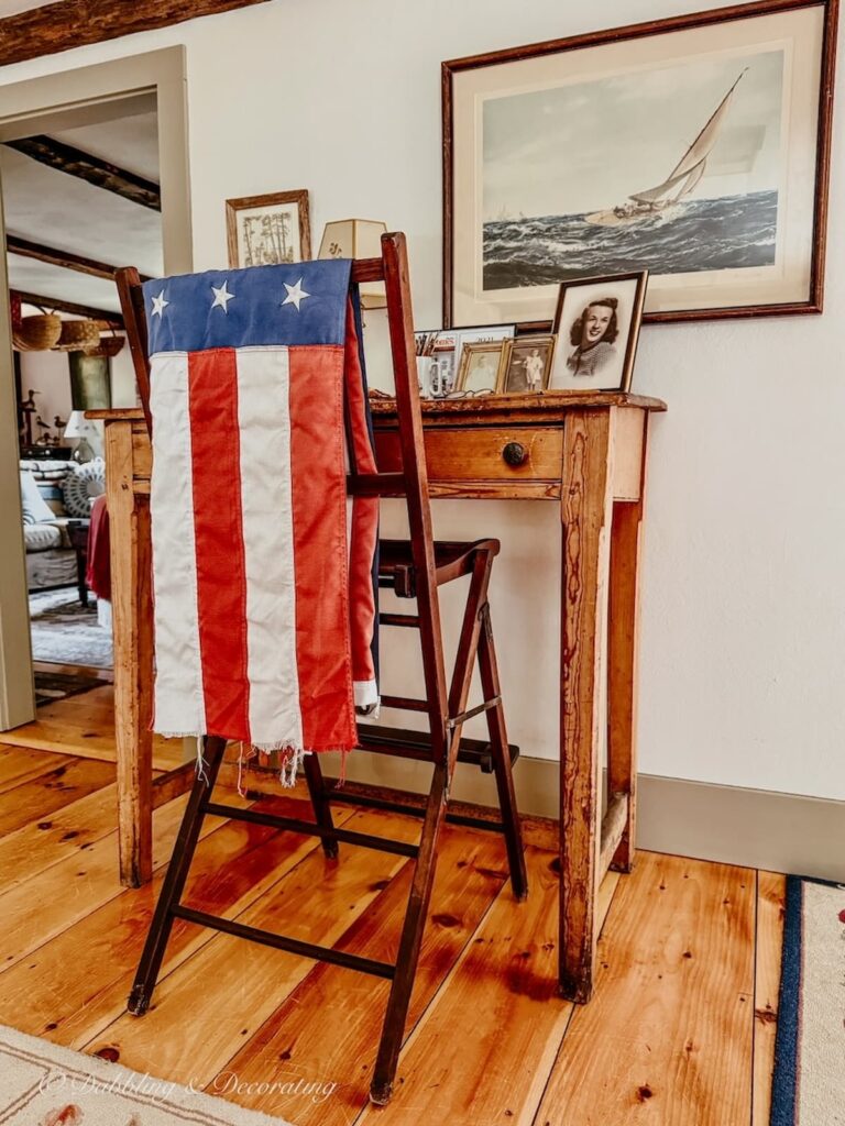 Old American flag draped over the back of a tall chair positioned at an antique desk in a coastal Maine home for vintage Americana decor ideas.
