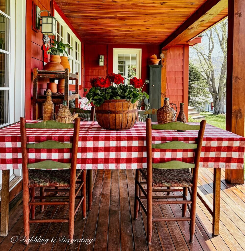 Covered porch with a table styled with red and white gingham tablecloth and red geranium centerpiece in wooden round basket for vintage Americana decor ideas outdoors.