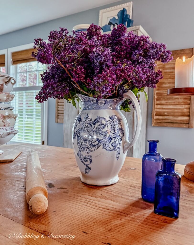 Vintage purple and white transferware pitcher filled a bouquet of lilacs on a wooden table paired with two small purple bottles.
