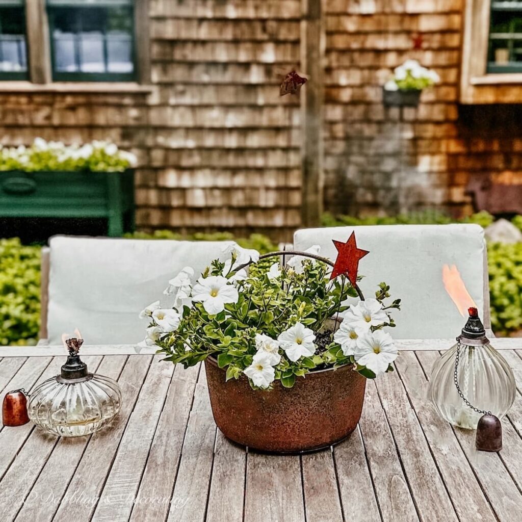 Antique wrought iron rustic pot filled with white petunias on teak table in front of New England cedar shake home for vintage Americana decor ideas.
