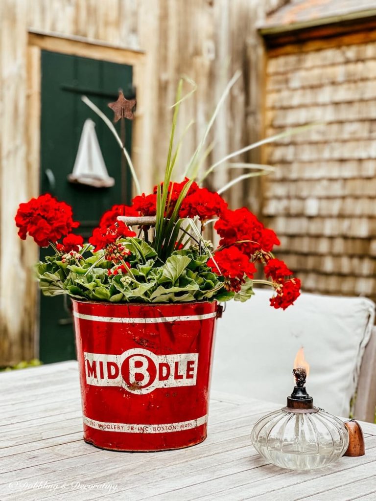 Vintage red bucket with red geranium arrangement on table.