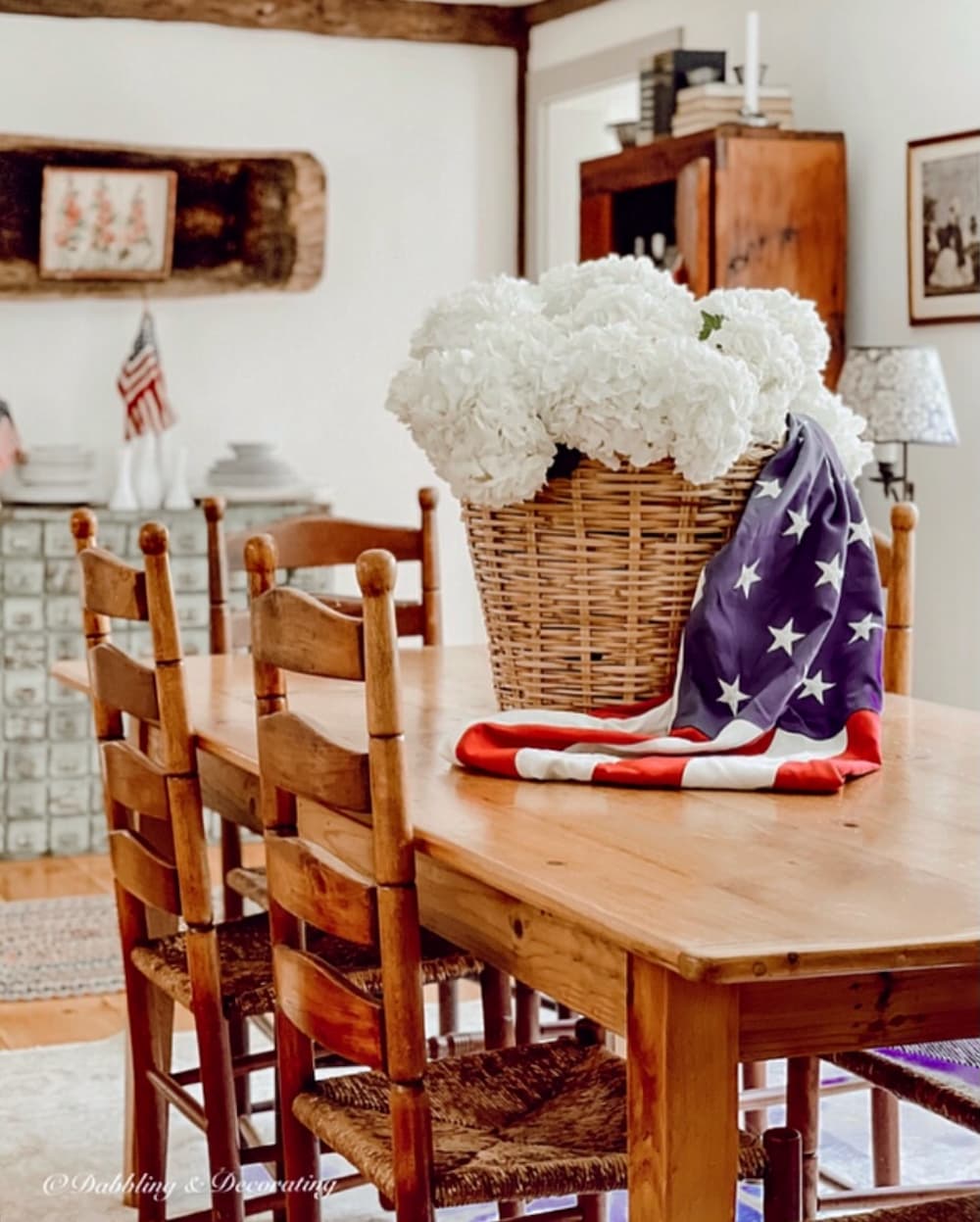 Antique styled dining room in Maine home styled with vintage Americana decor ideas with basket of white hydrangeas, USA draped flag as table centerpiece.