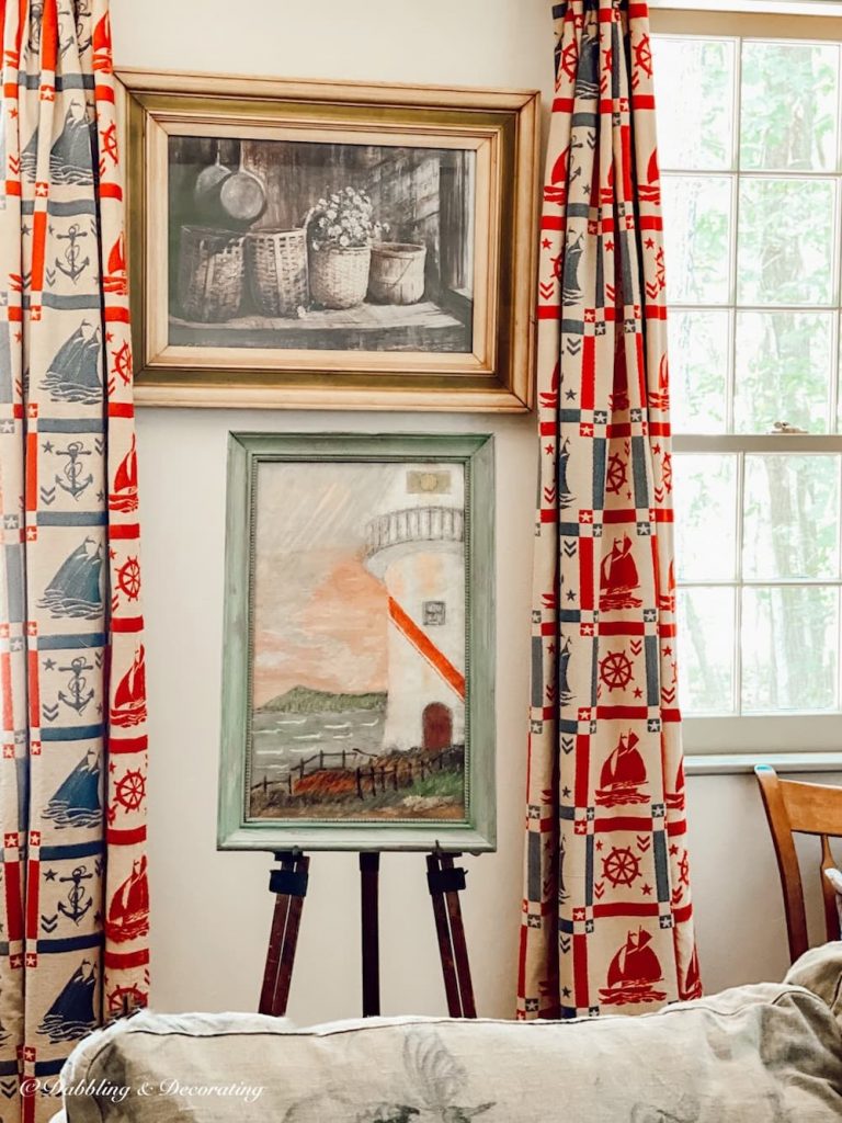 An antique easel with lighthouse painting next to living room windows with nautical style curtains.