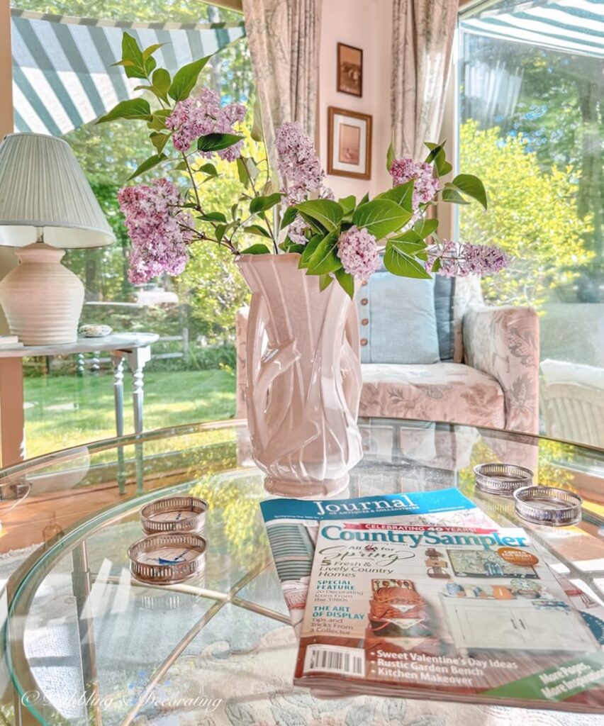 Vintage white McCoy vase with lilacs on glass coffee table with Country Sampler Magazine and Antique Journal displayed in sunroom.