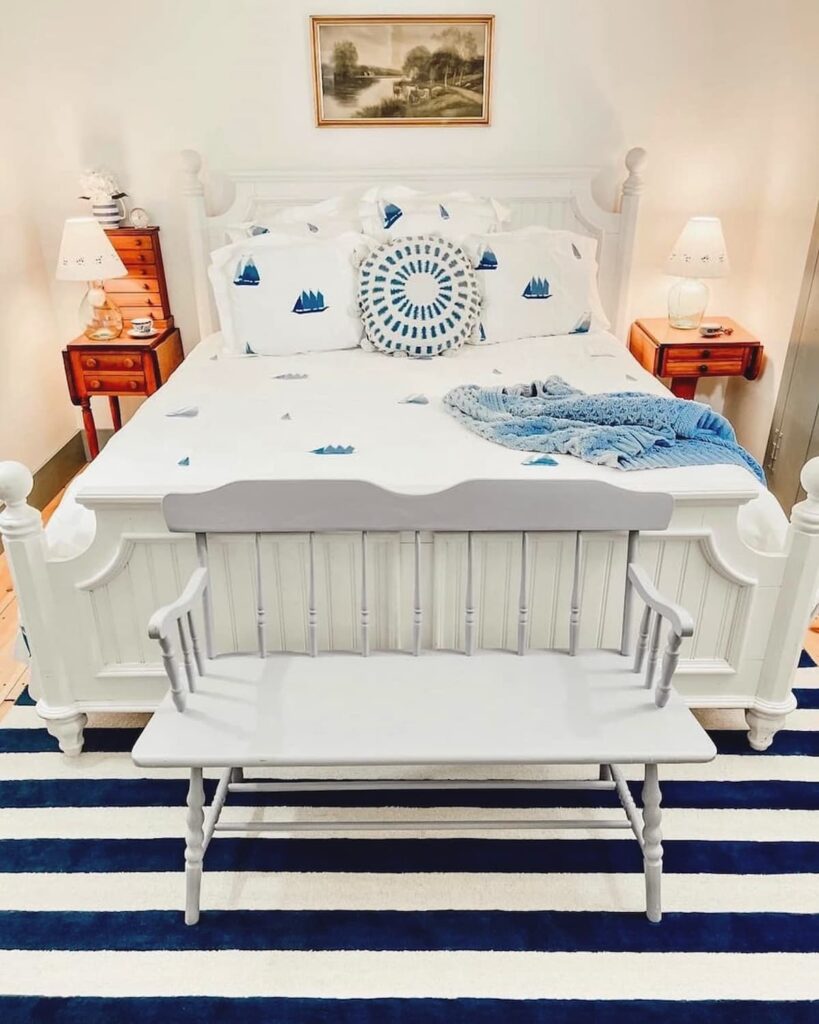 Coastal bedding ideas in vintage master bedroom with Schweitzer Linen Winward Collection in stark white and blue embroidered sailboats layered with pillows and navy striped rug.