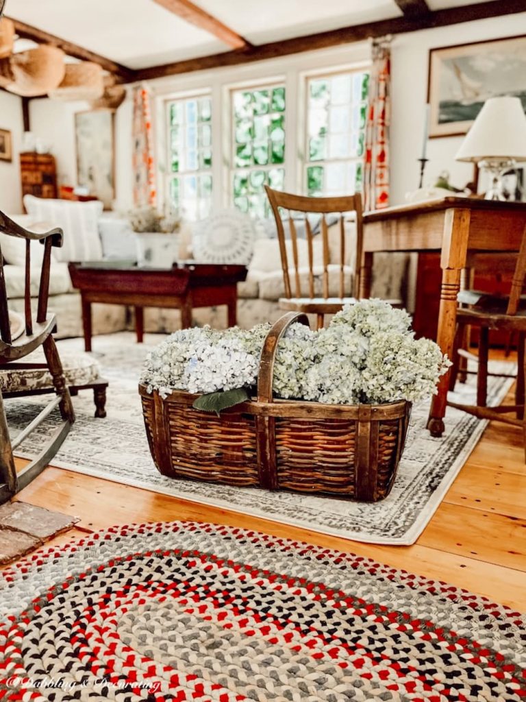 An antique styled living room with a vintage flower arrangement including wicker basket and blue hydrangeas on the floor and a rocking chair.