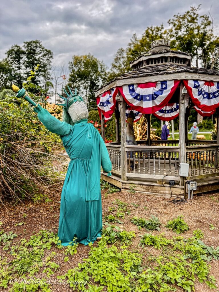 Lady Liberty as a Funny scarecrow next to a Gazebo covered in American flag bunting in fall decorations outdoors.