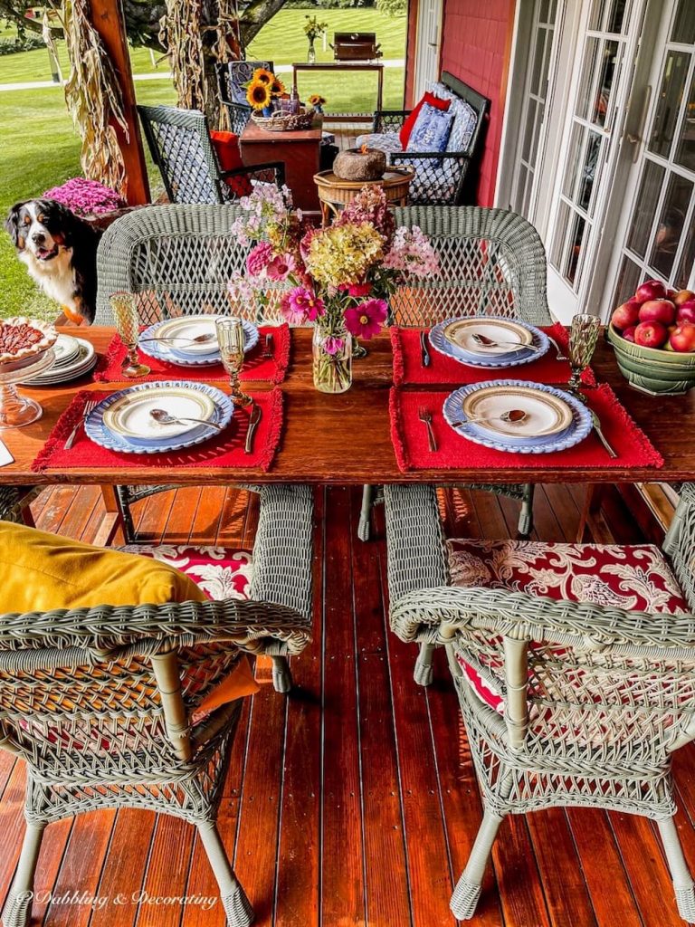 Covered fall decorated porch with vintage wallpaper table styled for dining and wicker chairs.