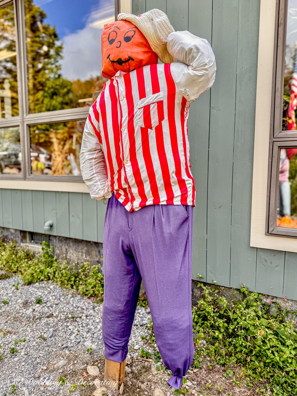 Funny scarecrow with red and white striped shirt, purple pants and orange face smiling.