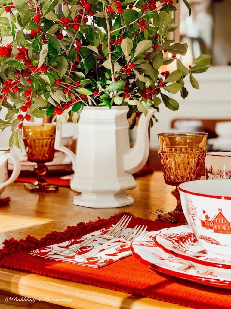Vintage white ceramic tea pot with winterberries centerpiece with red and white dinnerware for the holidays on table.