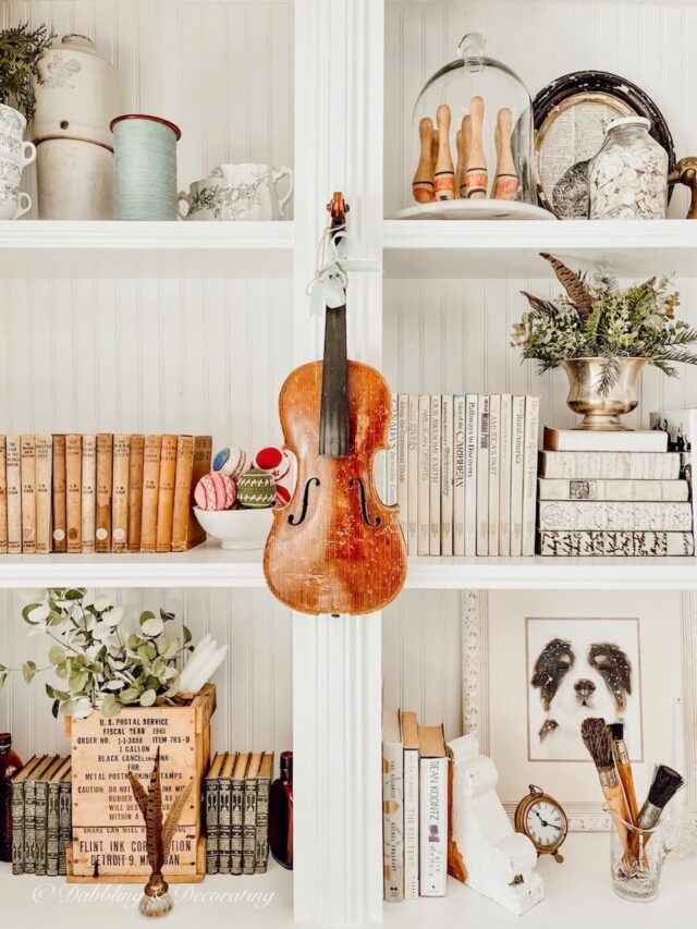 A vintage white bookcase adorns the living space, showcasing a delicate violin elegantly suspended from its shelves.