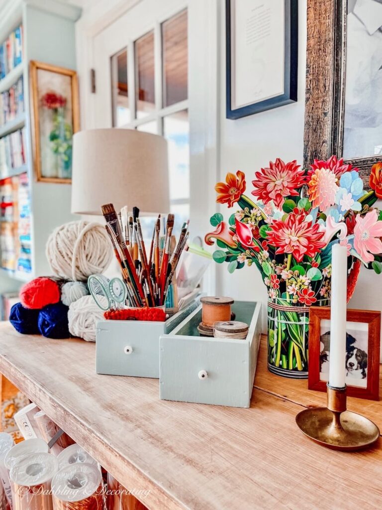 Vintage wallpaper table in craft room decorated with colorful eclectic craft supplies and lighting.