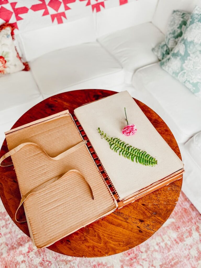 Vintage flower press opened up on table with a green fern leaf and pink flower on craft room table.
