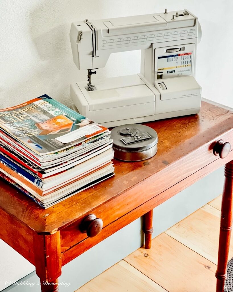 Stack of vintage Early American Life magazines on antique table with sewing machine in bedroom.