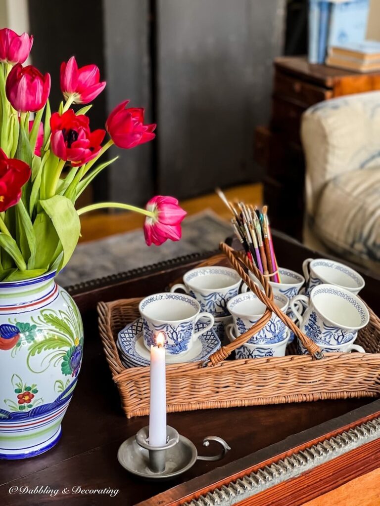 Italian ceramic vase with pink tulip arrangements on antique coffee table in living room with basket of teacups and lit pewter candle.