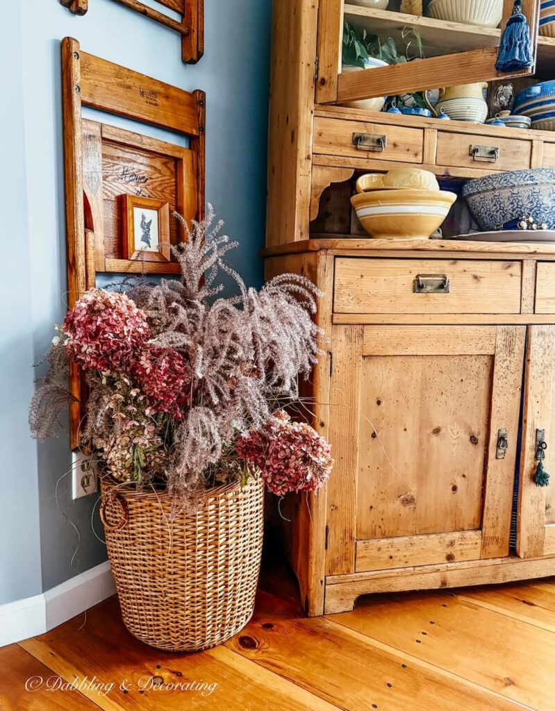 Pink and purple fall hydrangeas in a basket on the floor next to a vintage wooden pine cupboard in fall home decor ideas.