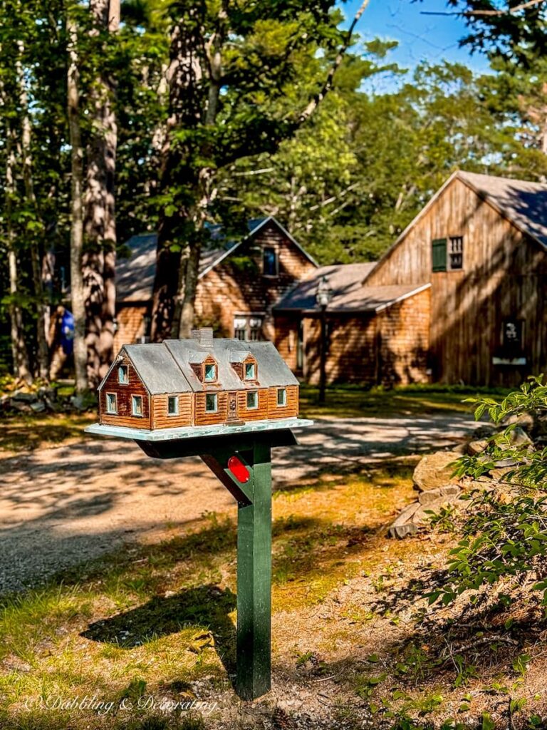 Unique mailbox house styled with cedar shakes on green pole in front of cedar shakes Maine home.