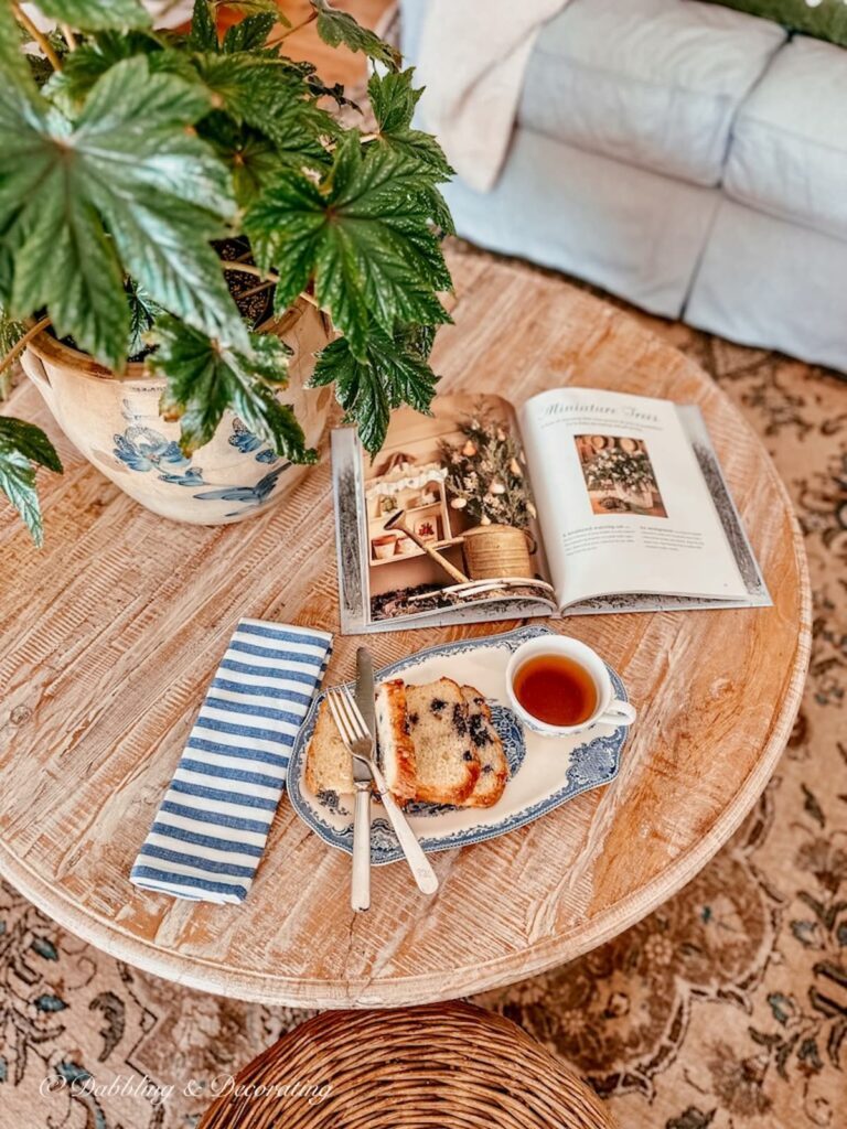 Round coffee table with green plant in vintage crocks, Johnson brothers tea and toast plate with blueberry cake and tea and Christmas with country living book on table.