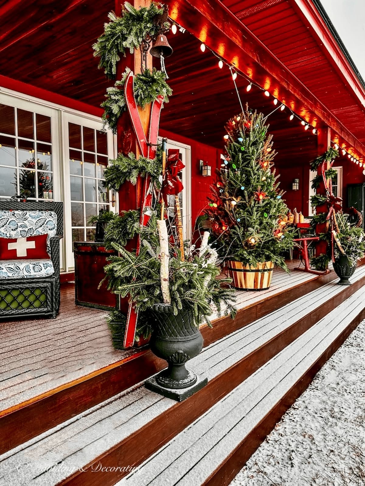 Vintage Christmas decor on porch of red house. Colorful lights on Christmas tree in whisky barrel, vintage red skis with evergreens and a Nordic cross pillow with snow. Vintage Skis for Decoration.