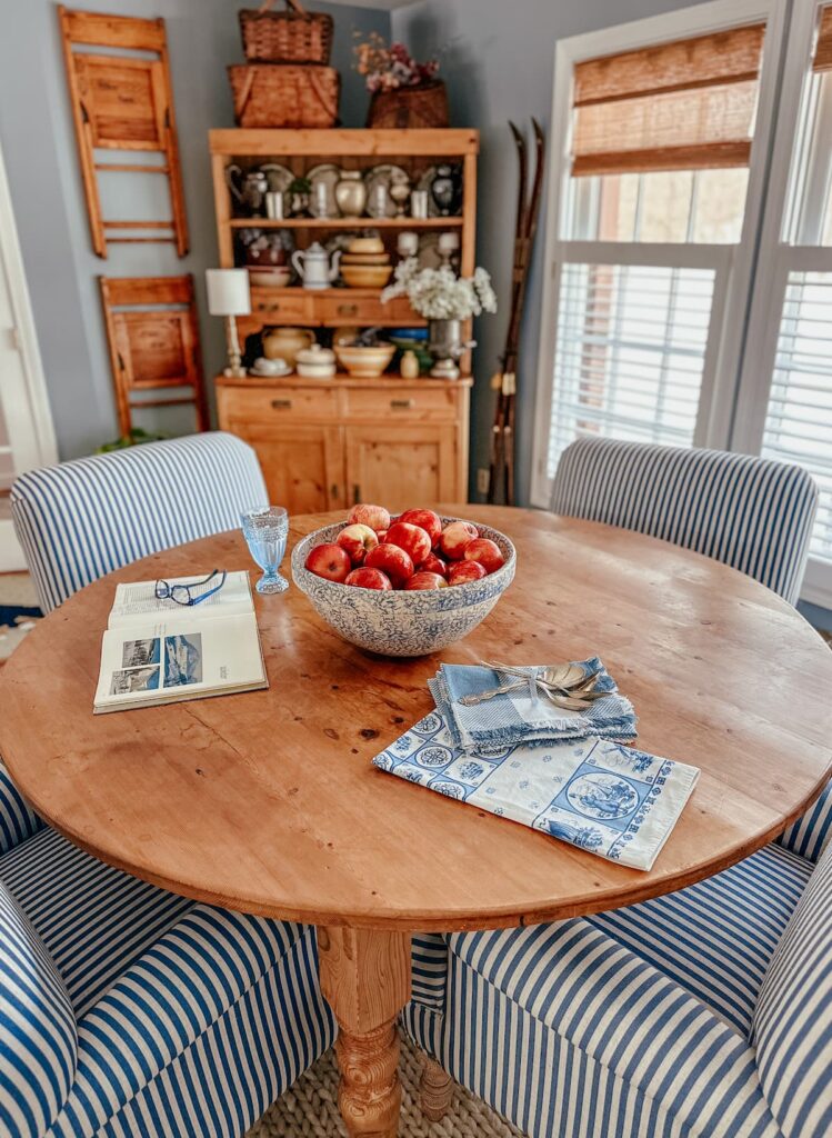Round vs. rectangular dining table with bowl of apples, blue and white striped chairs, vintage hutch and bamboo window shades on windows.