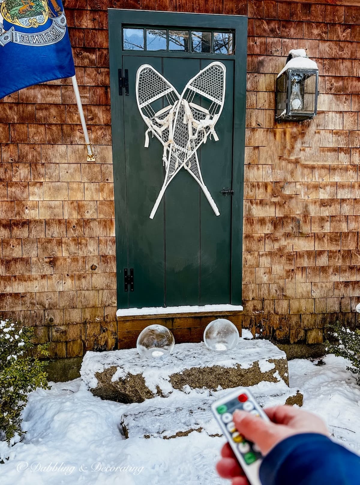 Cedar shake home with dark green front door with white snowshoes with two DIY ice lanterns on door step and Maine flag.