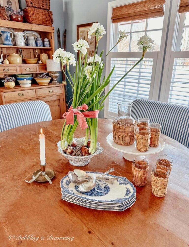 Paperwhite arrangements and vintage dishes with candle on pine dining room table, hutch and bamboo shades and shutters on windows.