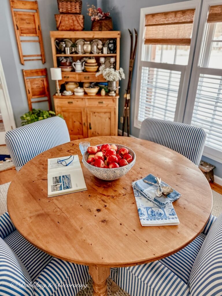 Dining room with large round table with bowl of apples, blue and white striped chairs, vintage hutch and windows with no-drill woven shades.