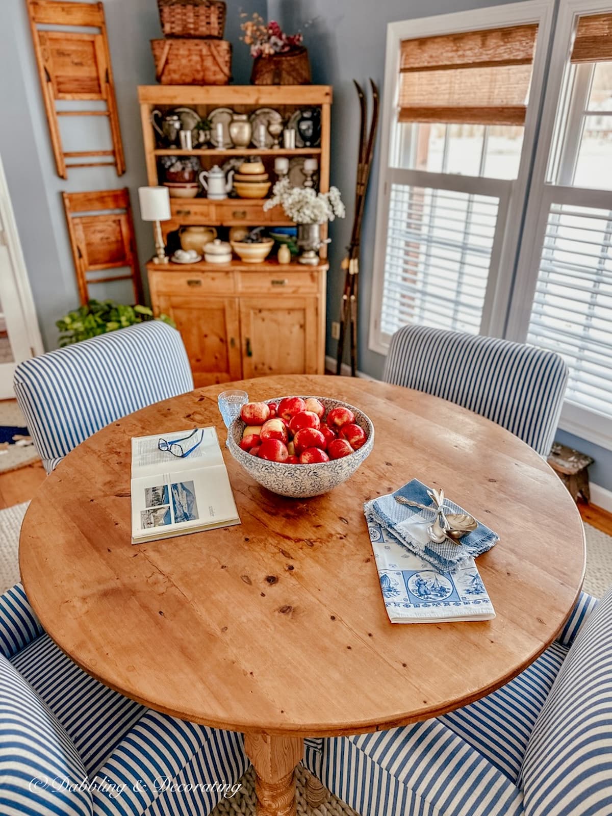 Dining room with large round table with bowl of apples, blue and white striped chairs, vintage hutch and windows with blinds and bamboo shades.