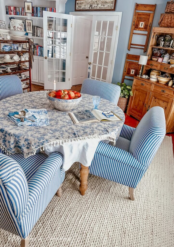 Vintage style dining room with blue and white chairs around round table on white rug with vintage wall decor and hutch.