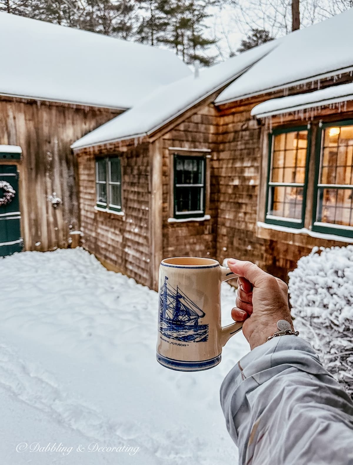 Blue and tan Delft mug in hand with coffee toasting to a cozy snow day outside of cedar shake siding home.