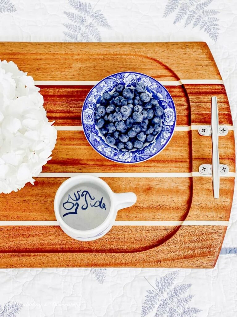 Wooden serving tray with low tide mug, bowl of blueberries and white hydrangeas with beautiful silver boat handles.