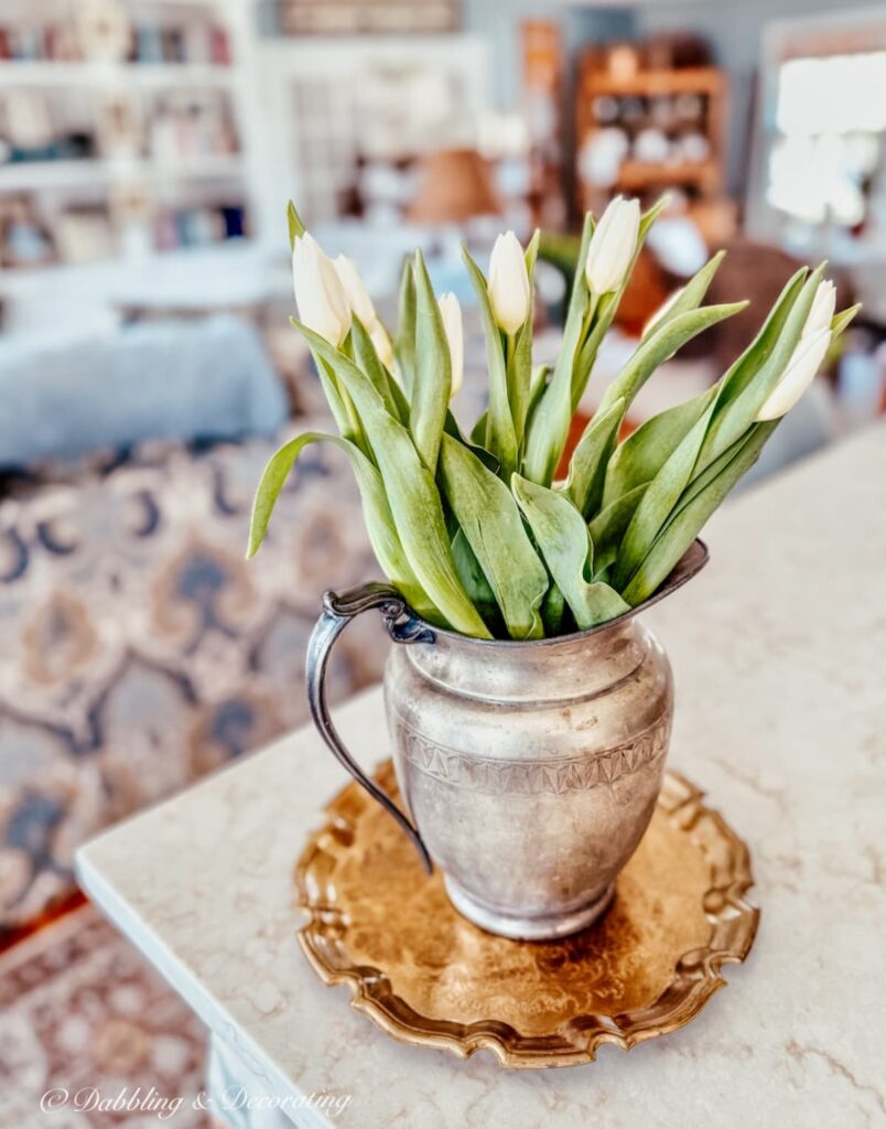 Silver vintage pitcher with white tulips on medium gold vintage tray on vintage open kitchen counter.