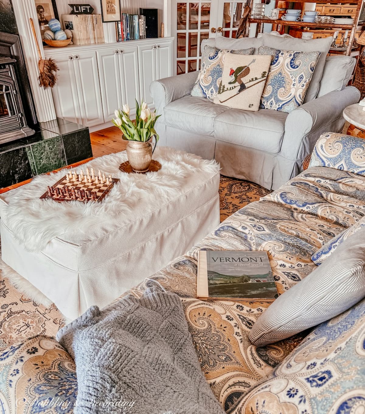 Living room ottoman with white sheepskin, ottoman tray with pitcher of white tulips, chess board game and blue and white paisley furnishings in front of pellet stove.