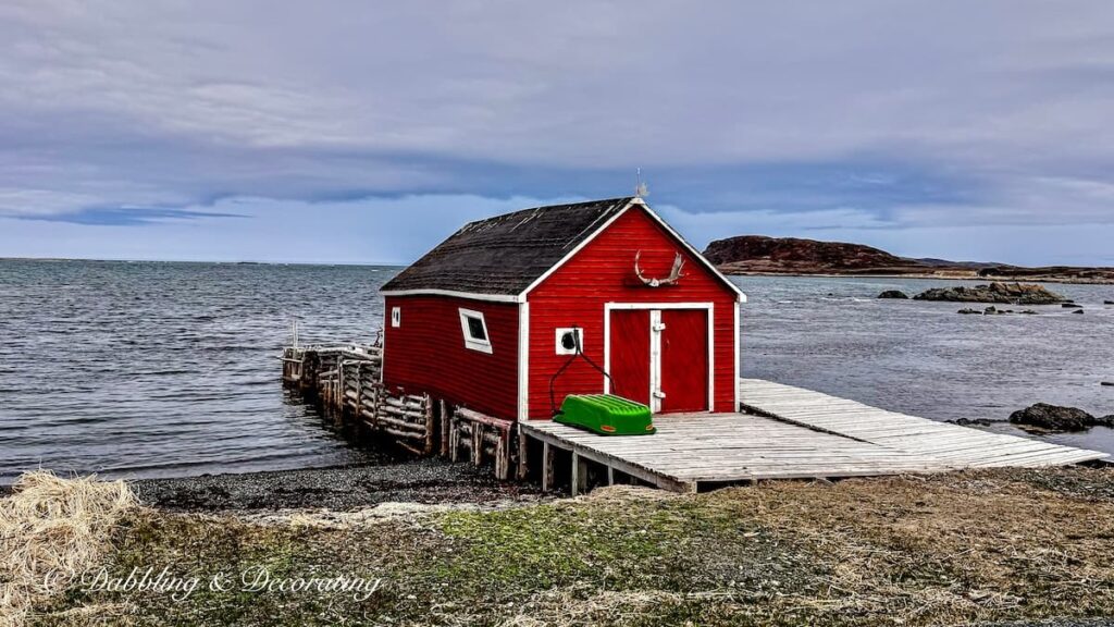Red fishing house with green boat standing alone on Newfoundland waters as inspiration for coastal cottage décor ideas.