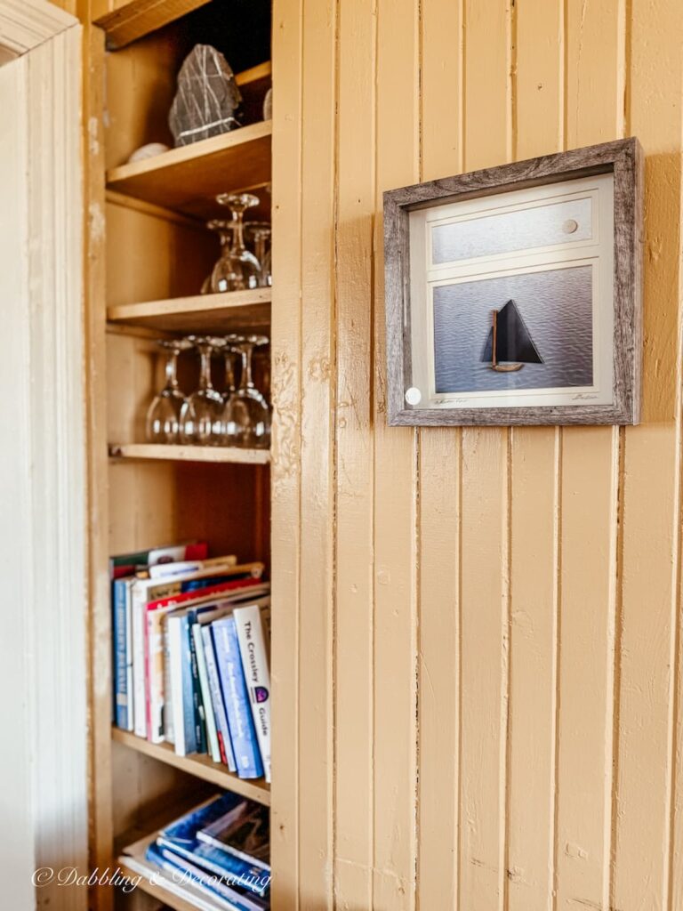 Coastal cottage decor shelves styled with books and glassware and a driftwood framed print on wall in lighthouse.