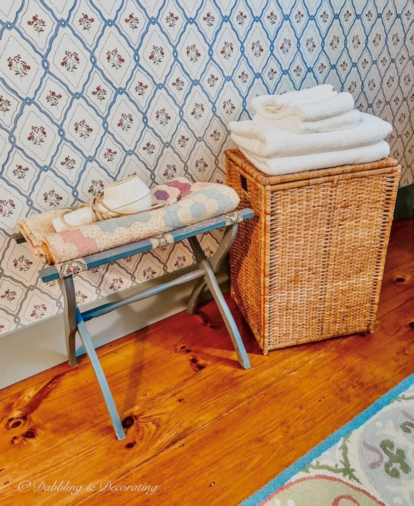 Wicker hamper with white towels against wallpapered wall with vintage luggage rack, quilt, and blow dryer in bedroom.