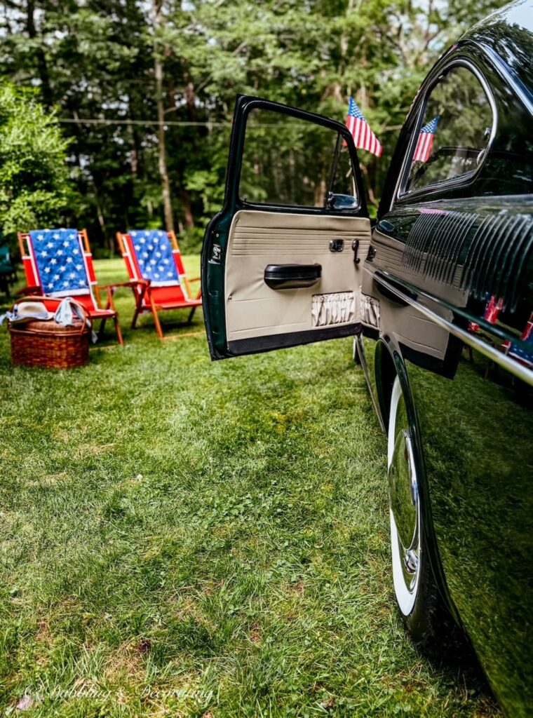Vintage car with door open styled next to red chairs with red, white, and blue towels and picnic basket in backyard.