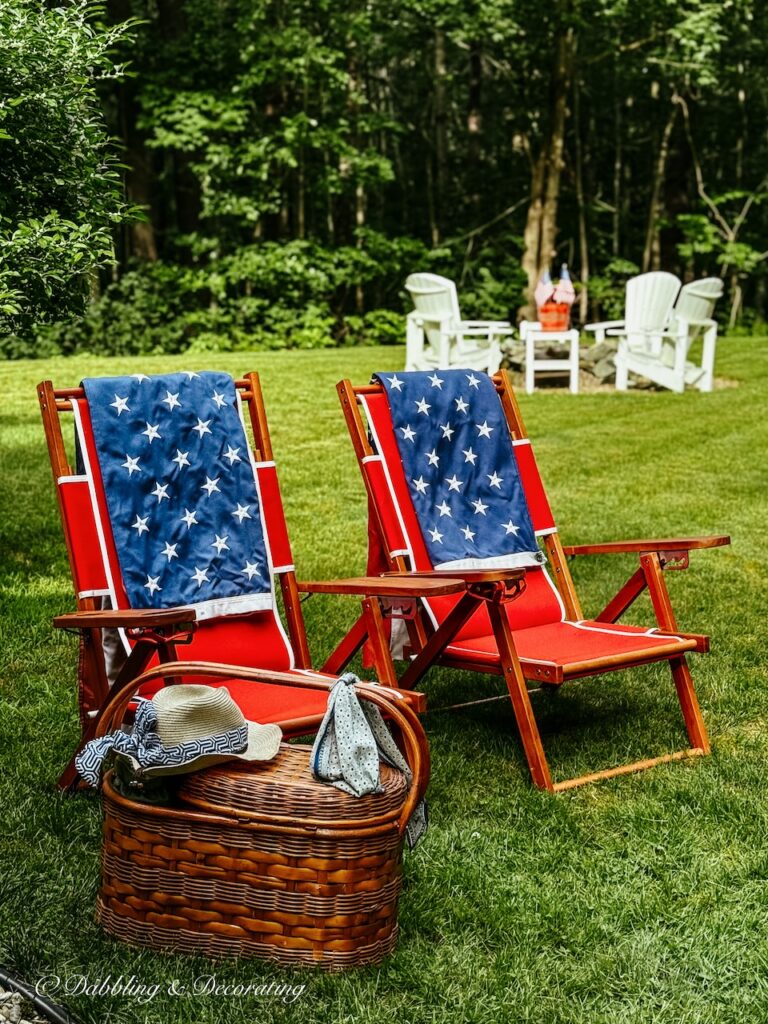 Red teak outdoor folding chairs in backyard with blue and white towels and picnic basket in backyard for 4th of July.