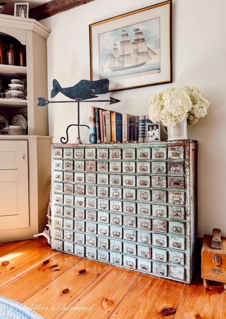 Antique apothecary cabinet with wrought iron weathervane, sailboat picture, books and a bouquet of white hydrangeas in old home.