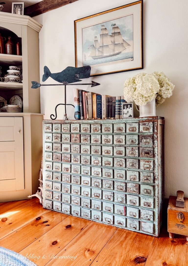 Antique apothecary cabinet with wrought iron weathervane, sailboat picture, books and a bouquet of white hydrangeas in old home.