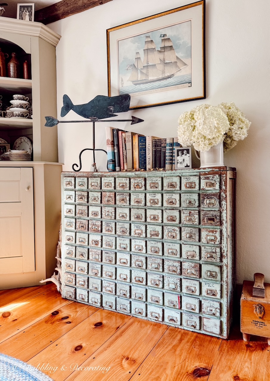 Antique apothecary cabinet with wrought iron weathervane, sailboat picture, books and a bouquet of white hydrangeas in old home.